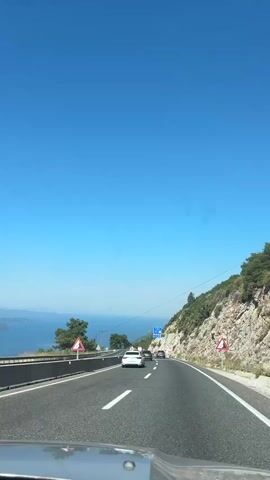 The video shows a car driving on a highway alongside a body of water and a rocky, tree-covered hillside under a clear blue sky. Several other cars are visible on the road ahead. Traffic signs warning of a curve are present on the right side of the road. The view is from the perspective of someone inside the driving vehicle, looking forward through the windshield.