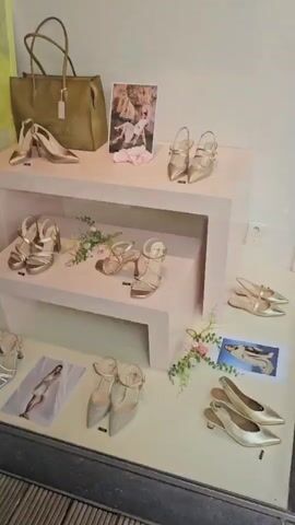 The video pans across a store window display. The display features a variety of women's shoes, handbags, and clothing. A large painting of a woman with a yellow hat and earrings is visible on the left side of the display. In the center, there are several pink shelves displaying gold and silver heels and sandals. A gold handbag sits on the top shelf. To the right, a framed picture of two women in white dresses is displayed. On the floor, more shoes are arranged, along with framed images of models wearing shoes. A mannequin dressed in white pants and a patterned top is visible behind the display. In front of the store, a large white planter with pink hydrangeas is placed. The store entrance is visible in the background, with a glass door and a doormat. A wreath and the number "40" are visible on the glass door. The video continues to show more of the store's interior, with racks of clothing and additional displays. The floor in front of the window is made of wooden planks.