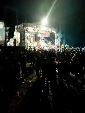 This media file captures a lively outdoor concert or festival event at night, likely in Krebet, Indonesia. A stage, supported by a large metal truss structure, is brightly lit with stage lights and spotlights, creating beams that cut through the darkness and illuminate the crowd. On the stage, a band or musical ensemble is performing. Several individuals are visible, some playing instruments like keyboards and guitars, while others appear to be singers or performers.

In front of the stage, a large crowd of people is gathered, most of them silhouetted against the stage lights. The audience appears to be actively engaged with the performance, with many individuals raising their hands, dancing, and enjoying the music. The overall atmosphere seems energetic and celebratory.

Visible text includes signage on the stage, with one prominent banner displaying "KREBET" in large letters, confirming the location. Another banner on the side of the stage has vertical text that is partially obscured but appears to say "VOYAGE MUSIC". A screen is also visible to the left of the stage, displaying some form of visual content, possibly related to the performance or advertisements.

The time of day is clearly night, indicated by the darkness of the sky and the reliance on artificial lighting. No specific weather conditions are evident, but the event is taking place outdoors, suggesting it is likely dry. The ground appears to be uneven and possibly dirt or gravel, typical of an outdoor venue. The emotions conveyed are those of excitement, joy, and communal enjoyment of a live musical performance.