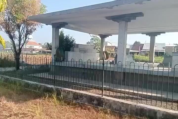 The video shows a fenced-off area with several stone structures, possibly tombs, covered by a concrete roof supported by columns. The area appears to be outdoors, with dry grass and vegetation visible in the foreground and background. Houses and trees can be seen in the distance, suggesting a rural or semi-rural setting. The sky is clear and the lighting indicates daytime.