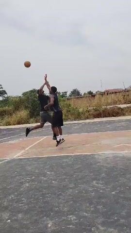 Two men are playing basketball on an outdoor court. The court is marked with white lines, but the paint is faded. The men are wearing casual athletic clothing. One man shoots a basketball, but it misses the hoop. There are some trees and tall grass in the background and a cloudy sky overhead.