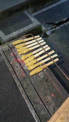A charcoal grill with eight yellow satay skewers rests on a textured, dark surface. The grill is rectangular, made of weathered metal, and stands on a simple metal frame. Hot coals glow red beneath the grate, indicating recent use. The skewers are made of light-colored wood and hold oblong pieces of food that appear to be marinating in yellow sauce. A pair of white sneakers with reflective strips can be seen in the background, suggesting a person is nearby. The background is dark and includes grey paving stones and indistinct shapes. The overall lighting indicates the scene is set outdoors at dusk or at night.