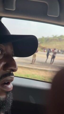 From inside a vehicle, we see a view through the side window of a group of people gathered along a road. Some are standing on the road itself, while others are on the grassy verge. The people are diverse in appearance, with varying clothing styles. Cars and motorcycles are also visible on the road. In the background, there is a hilly landscape dotted with trees and some buildings. The sky is overcast. The video appears to be shot from the perspective of someone inside the vehicle, with parts of the car's interior and the person holding the camera visible in the foreground.