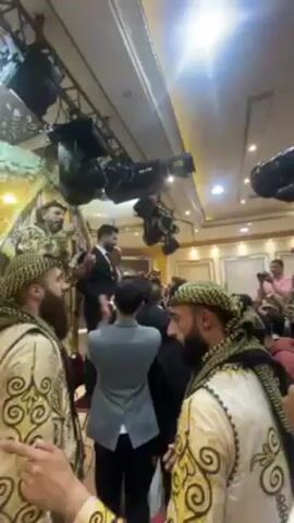 The video captures a lively celebration, possibly a wedding reception, inside a large hall with cream-colored walls and a high, white ceiling adorned with recessed lighting and an elaborate chandelier. The room is filled with men, most of whom appear to be Middle Eastern. Some are wearing traditional attire with patterned headscarves and embroidered vests.

The focus of the video is a man in a dark suit standing on what appears to be a raised platform, surrounded by celebrants. Several people are holding cameras and cell phones, recording the event. One man is holding a professional camera mounted on a gimbal. He lifts it above the crowd, attempting to get a better shot of the festivities. Overhead, complex lighting and sound equipment is rigged to the ceiling, adding to the atmosphere of a professionally organized event. The overall impression is of a joyful and well-documented celebration.