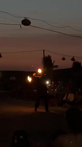 A person is performing a fire-spinning act at dusk. They are in a sandy area with several other people watching them. The performer is juggling two flaming objects, possibly torches or poi. The sky is a gradient of orange and dark blue, indicating the sun has set. There are lights strung across the scene, some of which are illuminating small tables where people are seated. Palm trees are visible in the background, and there are also structures that appear to be tents or shaded areas. The overall atmosphere seems to be festive and outdoor-oriented.