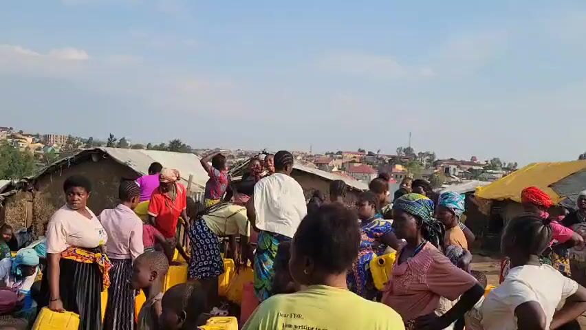 A crowd of people, mostly women and children, are gathered outdoors. They appear to be in a village or informal settlement, with simple structures visible in the background. Many individuals are holding yellow plastic containers, suggesting they are collecting water or some other commodity. Some people are interacting with each other, while others are looking in different directions. The overall atmosphere seems to be one of activity and perhaps a communal gathering. The sky is visible above, indicating it is daytime.