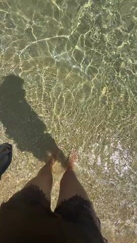 The video begins with a close-up shot of a person's legs wading into clear, shallow water. The water is illuminated by sunlight, creating shimmering patterns on the sandy bottom. The camera then pans up to reveal a wider view of a beach scene. The sky is a clear, bright blue, with a few wispy clouds. The ocean water is turquoise and crystal clear, with gentle waves lapping the shore. In the distance, a sandy beach is lined with umbrellas and beachgoers. The video ends with a view of the open sea.
