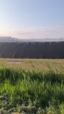 A drone or camera moves forward, capturing a view of a large, dark, terraced structure resembling a landfill or an industrial berm. In the distance, the sky is clear and blue with a few wispy clouds. Beyond the berm, several white buildings or structures are visible. In the foreground, green grass and weeds line the bottom of the frame. Some of the grass appears to have been recently cut. The overall scene suggests an outdoor, possibly rural or industrial, environment. There are no visible people or animals in the video.