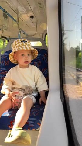 A young child wearing a beige t-shirt and patterned bucket hat sits on a bus seat, looking out the window. The child's tongue is sticking out slightly. The bus interior is visible, with blue and white seats and overhead handrails. Outside the window, a grassy landscape and train tracks are visible. The sun is shining, casting shadows and highlights.