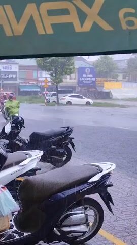 The video shows a person on a white scooter wearing a green poncho and a purple helmet riding through heavy rain. The scooter is splashing water as it moves. In the background, there are other vehicles, buildings, and trees. The scene appears to be a street in a town or city.