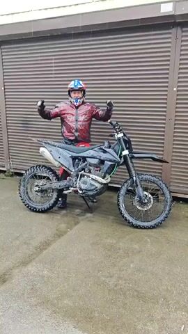 A man in a motorcycle helmet, a shiny red jacket, black gloves, and black pants stands next to a black dirt bike. He raises his arms in the air and makes a peace sign with one hand, smiling. The motorcycle is dirty, and the ground is wet, suggesting recent use in muddy conditions. The background consists of a metal roll-up door, likely a garage. Rain is falling.