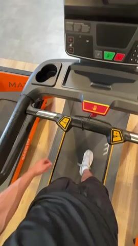 The video begins with a close-up shot of a person's legs and feet on a treadmill, with their right foot in mid-stride. The camera then pans up to show the control panel of the treadmill, displaying a time of 0:46 and other workout metrics. The scene appears to be a gym, with other treadmills and gym equipment visible in the background. There are other people in the gym, one of whom is walking away from the camera in the distance. The treadmill is a MATRIX brand, and the gym seems to be called MAGIC FIT. A sign in the background indicates "VESTIAIRES HOMMES," which translates to "Men's Changing Rooms." The overall environment is brightly lit and appears to be a modern fitness facility.