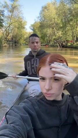 A selfie-style video shows two people on a canoe ride. The person in front is a woman with auburn hair, wearing a gray top, and has tattoos on her hand. She makes a "V" hand gesture at the camera. Behind her is a man with multiple facial and neck tattoos, wearing a dark hooded sweatshirt. He holds a paddle and smiles. The canoe is moving on a narrow waterway with muddy water. Trees line the banks on either side. In the background, wooden docks and structures are visible. The sky is clear. The camera appears to be held steady, capturing the motion of the canoe.