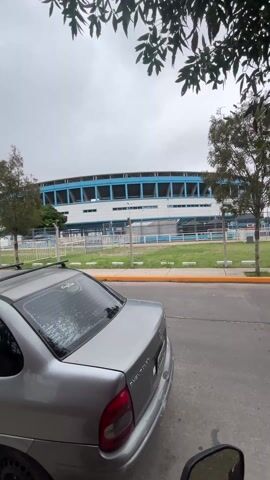 The video shows a street scene with several parked vehicles next to a stadium on an overcast day. The camera pans slightly from a gray sedan with a roof rack, revealing a white Volkswagen Amarok pickup truck and a patterned white van parked ahead. A lamppost displays an advertisement featuring a man and child. The stadium, painted in blue and white, is visible across a chain-link fence. Trees line the street and the sky is uniformly gray.
