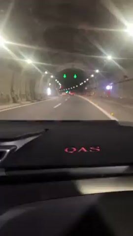 A car is driving through a well-lit tunnel. The interior of the car is visible, with the word "QAS" embroidered in red on the dashboard. The tunnel has lights along the walls and ceiling, and there are green arrows pointing downwards above the road. There are other cars ahead in the tunnel. A person can be heard speaking.