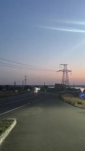 The video shows a road at dusk. The sky is a soft gradient of blue and pink. A large, metallic power line tower stands on the right side of the frame. Several other power line towers are visible in the distance. There are cars on the road, their headlights illuminating the asphalt. A few buildings and trees can be seen on the left side of the road. The overall impression is one of a rural or semi-rural setting at the end of the day.