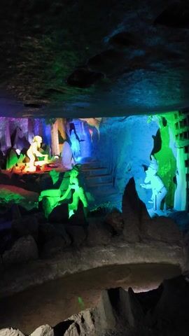 The video depicts a dark, narrow space resembling a cave or passage, illuminated by bright, colorful lights. The colors shift from blue to green to purple and red. There appear to be stone structures on either side of the passageway, and in the distance, there are vague shapes that could be figures or statues, also illuminated by the colorful lights. The overall scene is surreal and artificially lit.