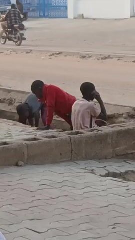 Three young people are crouched near a concrete curb with their hands inside what looks like a small drainage ditch. The person on the left is wearing a gray shirt and burgundy pants and is bending over. The person in the middle is wearing a red shirt and has their arm extended into the ditch. The person on the right is wearing a pink shirt and jeans and appears to be speaking. In the background, people are on foot and riding vehicles. A person is standing behind a pushcart with goods, and another person is riding a bicycle. The area is dusty and appears to be an urban street in a warm climate.