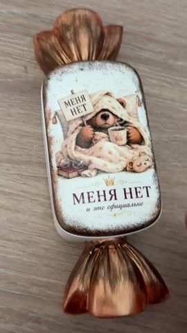 The video shows a person's hand holding a rectangular object shaped like a candy. The object has a white base with a candle inside and decorative copper-colored ends resembling candy wrappers. The front of the object features an illustration of a bear wrapped in a blanket, holding a mug and a sign that says "MENYA NET" (I am not here). Below the illustration, the text "MENYA NET and it's official" is written. The hand rotates the object, revealing the candle inside and then turning it back to show the illustration.