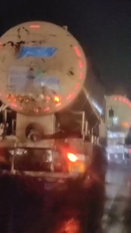 The video shows a nighttime scene with several large tanker trucks. The trucks appear to be white or light-colored and have reflective red and orange markings on their cylindrical tanks. The ground is wet, suggesting it may have been raining or is damp. The lighting is dim, and the headlights and taillights of the trucks are visible, casting reflections on the wet surface. Some trucks have visible writing on their tanks, but it is difficult to read due to the low light and image quality. The overall impression is of a highway or industrial area at night with several large vehicles present.