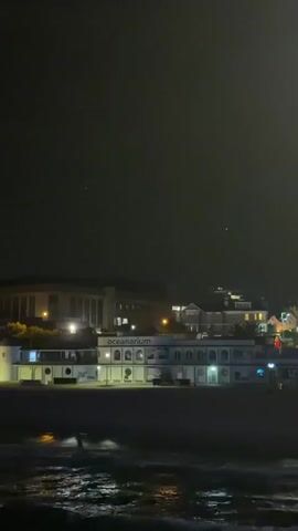 The video shows an oceanarium at night. The building is illuminated, and the ocean is in the foreground. The water is choppy, and there are lights reflecting on the surface. The sky is dark, and there are a few stars visible.