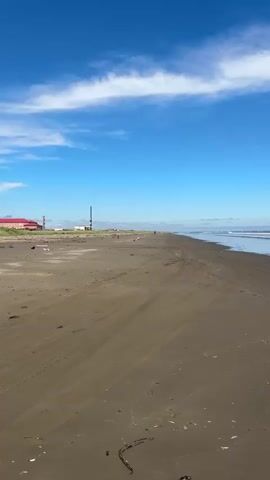 A wide shot of a dark sand beach under a bright blue sky with scattered white clouds. The tide is out, leaving a flat, wet sand surface with gentle wave patterns. In the distance, the ocean meets the horizon, with small waves breaking. On the left side of the frame, some distant buildings and industrial structures are visible, along with a few faint figures walking on the beach. The overall atmosphere is serene and natural.