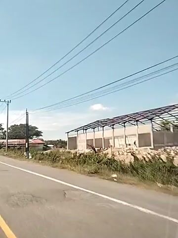 A video taken from a moving vehicle shows a paved road with a yellow line on the left side. Overhead power lines run parallel to the road, supported by utility poles. On the right side of the road, a partially constructed building with a metal frame is visible. Further down the road, there are some small roadside structures and trees. On the left side of the road, there are more trees and some low-lying buildings. A dark-colored van is seen approaching from the left. The sky is clear and blue with some clouds. The overall scene suggests a rural or developing area with ongoing construction.