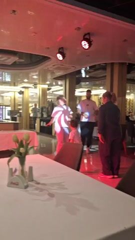 The video shows a brightly lit restaurant with people moving around. The camera is focused on a table with a white tablecloth and a vase of yellow tulips. The ceiling has decorative lighting that changes color, including red, green, and purple. Several people are standing in the background. A child runs around the group. A buffet area is visible. The flooring appears to be light-colored tile. The overall atmosphere seems lively and energetic.