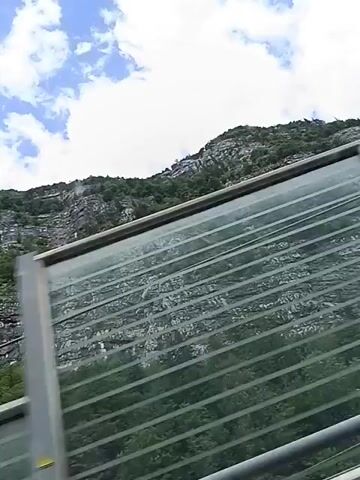 This is a first-person perspective video of a person traveling, presumably in a vehicle, as indicated by the motion blur and the presence of what appears to be a sound barrier or guardrail in the foreground. The barrier is constructed of metal framing and translucent panels, some of which are ribbed or textured. Behind the barrier, a steep, rocky mountain face covered in lush green vegetation dominates the scene. The sky above is a bright blue with scattered white, puffy clouds. The video captures a sense of movement as the scenery passes by, with the mountain appearing to shift and change perspective slightly. The camera pans slightly to the right as the video progresses.