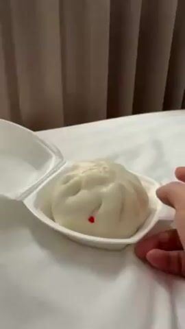 The video shows a white steamed bun inside a white styrofoam container on a white bed. A hand reaches into the frame, picks up the bun, and lifts it out of the container. The bun has a small red dot on it. A beige curtain is visible in the background.