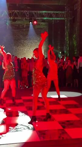 The video shows a group of people dancing on a checkered floor in a dimly lit venue. Three women in short, sparkly dresses are in the foreground, dancing in unison. They are wearing black boots. Red lights illuminate the scene, casting red shadows on the floor and walls. In the background, a crowd of people watch the dancers. There are stage lights and trusses visible above. The women appear to be performing a choreographed routine.