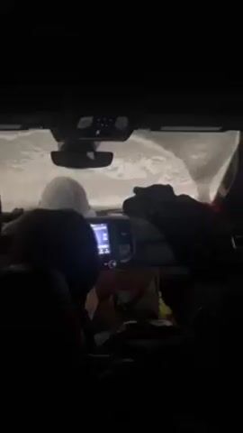 The camera is inside a dark vehicle with a white hardhat on the dashboard. The windshield is covered in what appears to be snow or ice, almost completely obscuring the view outside. The vehicle's interior lights are on and reflected in the rear view mirror. The center console features a screen, presumably the car's infotainment system. There is a person's head visible in the lower left corner of the frame. There are also dark objects, potentially clothing, obscuring much of the dashboard. The overall scene is dimly lit, with the primary light source being the interior car lights and some light from outside shining through the snow-covered windshield.