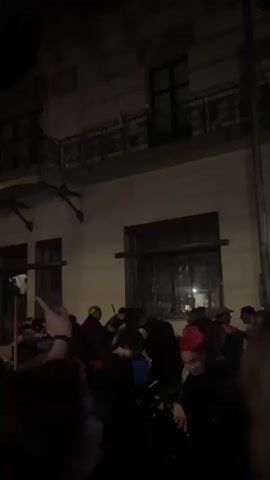 A dark, low-light video shows a crowd of people gathered outside a building at night. The building appears to be old with architectural details like balconies and large windows. The people are mostly dressed in dark clothing and many appear to be looking towards something or someone in the center of the group. Some individuals are visible in the foreground, including a woman with glasses. The atmosphere seems to be one of a social gathering or possibly an event. The exact nature of the gathering is unclear due to the low light conditions.