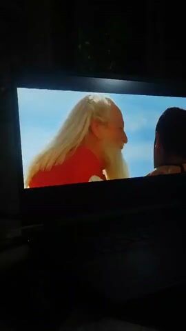 A laptop screen displays a scene from a movie. On the left side of the screen, an elderly man with long white hair and a white beard, wearing a red shirt, is looking towards the right. On the right side of the screen, a younger man with dark hair and a beard, wearing a brown and blue patterned vest over a white shirt, is looking towards the left. The background on the screen appears to be a bright, cloudy sky. The laptop is open and placed in a dimly lit room.