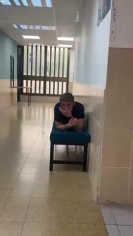A man in a navy blue shirt is lying prone on a small teal bench in a school hallway. The hallway has light tan tile floors and light colored walls, and overhead fluorescent lighting. There is a windowed wall at the end of the hall, and some posters are visible. The man is looking towards the camera and appears to be talking or gesturing.
