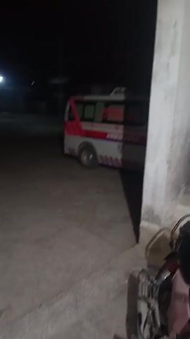The video shows an ambulance parked outside a building at night. The ambulance is white with red and blue stripes and the word "AMBULANCE" written on the side. The words "Rescue 1122" are written above the word "Ambulance." A motorcycle is parked next to the wall in the foreground. The area is dark.