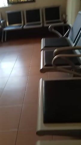 The video shows an interior space, possibly a waiting area. It features multiple rows of metal and upholstered chairs arranged along a wall beneath a window. The floor is tiled in light brown. There is an air conditioning unit mounted high on the wall. The camera pans to reveal a reception desk with a person sitting behind it, working on a computer.