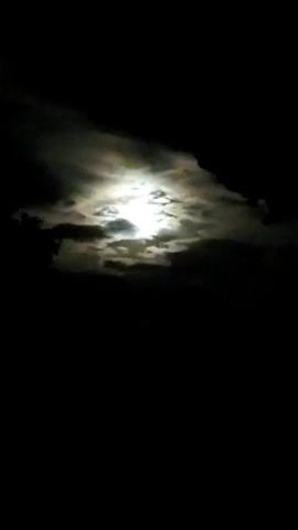 The video shows a full moon partially obscured by clouds at night. The moon is bright and illuminates the surrounding clouds, creating a dramatic effect. The foreground is completely dark, indicating that the shot was taken at night and the camera is pointed towards the sky.