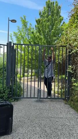 In Bron, France, a man is seen climbing over a tall, dark gray metal gate on a sunny day. He wears a light-colored short-sleeve shirt, dark pants, sneakers, sunglasses, and carries a dark shoulder bag. The gate is composed of numerous vertical and horizontal bars and appears to be closed.

At the beginning of the video, the man uses the gate's bars to hoist himself up, placing one foot on a lower horizontal bar and gripping the top with both hands. He then lifts his other leg, gradually maneuvering his body to sit atop the gate's structure. With apparent ease, he swings his legs over to the other side, successfully circumventing the barrier.

The scene is outdoors, featuring a gravel pathway leading up to the gate. Lush green trees and bushes are visible on both sides of and behind the gate, suggesting a natural or park-like environment. A lamppost stands to the left of the gate. The sky is clear and bright blue, indicating good weather. A black hardshell suitcase rests on the ground to the left, outside the gate, hinting at travel or movement. There is no visible text.