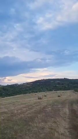 The video shows a landscape with rolling hills and fields. The sky is cloudy with patches of light suggesting a sunset or sunrise. There are hay bales scattered across the fields. In the distance, there are trees and some structures, possibly buildings or towers. The camera pans slowly across the scene.