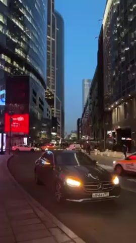 The video shows a dark-colored Mercedes-Benz car with its headlights on, parked on the side of a city street. The car’s license plate number is visible. The street curves slightly to the left. Tall, modern skyscrapers line both sides of the street, illuminated with lights. There are other vehicles on the street, including taxis. Pedestrians can be seen walking on the sidewalks. The sky is a clear, light blue color, suggesting it is dusk or early evening. Some buildings have digital billboards displaying advertisements.