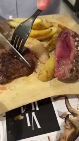 A person is cutting a rare steak on a wooden cutting board, with a black fork and knife. The steak is surrounded by cooked potatoes and sprinkled with salt. The steak is very rare, with a dark brown exterior and a deep red interior. The cutting board sits atop a printed newspaper.