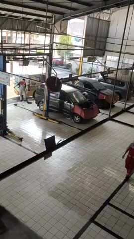 The video shows the interior of a car repair shop. The shop has a tiled floor and a metal roof. Several cars are parked in the shop. One car is on a car lift. Two mechanics are visible in the video. One mechanic is standing near a car, and the other is walking across the shop floor. The shop appears to be well-lit and organized.