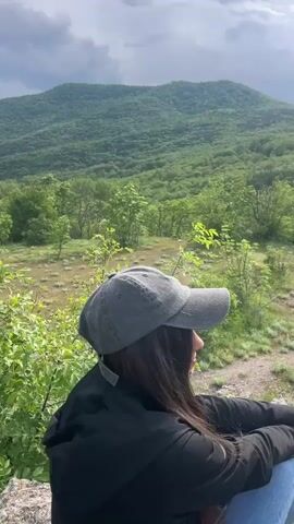 A medium shot captures a person wearing a gray cap and a black jacket looking out over a landscape, before the camera shifts to reveal the scene.  The landscape features a lush, green mountain ridge dominating the view, thickly covered with trees. In the foreground, there is a meadow-like area with sparse grass and small trees. A dirt path winds through the meadow. The sky is overcast with dark, heavy clouds, suggesting an impending storm or recent rainfall. The overall setting gives a sense of tranquility and natural beauty despite the cloudy weather.