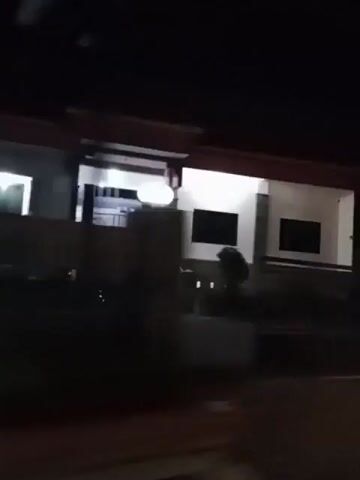 The video shows a dimly lit scene at night, possibly from a moving vehicle. The initial frame captures the exterior of a building with illuminated windows. The camera then pans to the right, revealing lush green foliage with a small structure featuring a red roof. Subsequent frames become increasingly dark, showing faint lights in the distance against a black background, suggesting the movement continues away from illuminated areas into a darker environment. The quality of the footage is poor, with motion blur throughout.
