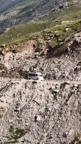 A white passenger van is seen driving on a narrow, unpaved mountain road. The road is rocky and appears to be undergoing some construction or has experienced a landslide, with loose rocks and debris on the side. The surrounding landscape is mountainous with steep, grassy slopes and jagged rock formations under a partly cloudy sky. The camera is positioned at a high angle, looking down on the road and the van.