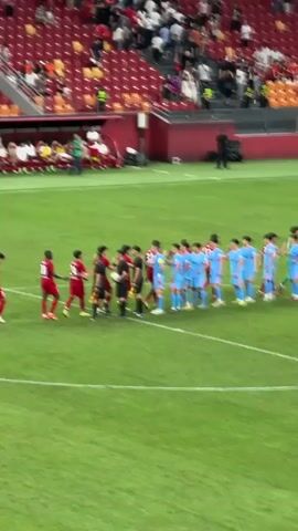 Here's a detailed description of the video:

The video captures a soccer match lineup on a bright, green field in a stadium. Two teams face each other, one dressed in red jerseys and the other in light blue. The teams shake hands as they line up. A referee, in a black uniform, stands nearby, managing the proceedings. The stadium seats behind them are partially filled with spectators. The overall atmosphere is one of anticipation and formality before a sporting event begins.