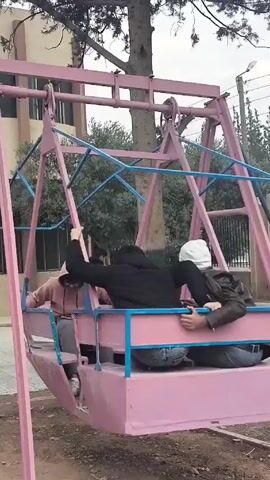 The video shows a group of people on a swing set in an outdoor environment, likely a park or schoolyard. The swing set is a large, rotating structure made of pink and blue metal. Two people are inside the enclosed part of the swing, while another person is on top, holding onto the frame. The swing is rotating, causing the people inside to move around. The setting includes trees, a building, and a partially visible parked red moped or scooter. The weather appears overcast.