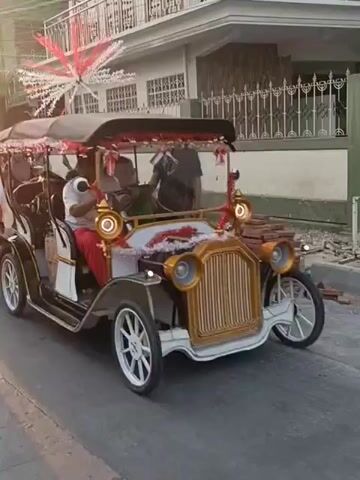 A decorated antique car, likely a Model T replica, is featured in what appears to be a parade or celebration. The car is white with gold accents and adorned with red and white streamers and flags, suggesting a patriotic theme, possibly for Indonesian Independence Day. It is carrying passengers and being driven on a street. Several other vehicles, including motorcycles and cars, are also present, along with a crowd of people walking and riding. The overall atmosphere suggests a festive outdoor event in a populated urban or semi-urban area. The sun is setting, casting long shadows and creating a warm, golden light.