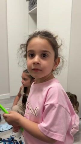 A young girl is the primary focus of the video, positioned in front of the camera. She has brown hair, tied up in a casual style, and is wearing a pink t-shirt with the text "Pretty & Cool THE ROOM" printed on it. The girl is seen smiling sweetly at the camera.

The background appears to be an indoor setting, possibly a room with white walls and a light-colored floor. A white shelf unit is visible in the back. Two other young girls are seated in the background; their presence is less emphasized as they are slightly out of focus. The first girl in the background is wearing a tan outfit. The other is wearing a pink t-shirt like the one the girl in front is wearing. The girl in front is holding what appears to be a green toy in her hands.